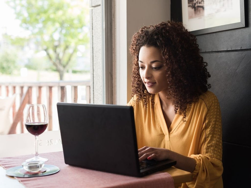 Woman in a mustard blouse types on a laptop at a cafe table with a glass of red wine, near a window in a calm setting.