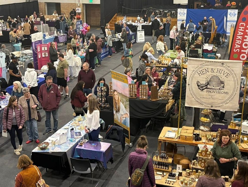 Indoor craft fair with people browsing various booths. Notable "Hen & Hive Provisions" sign. Colorful displays, lively atmosphere.