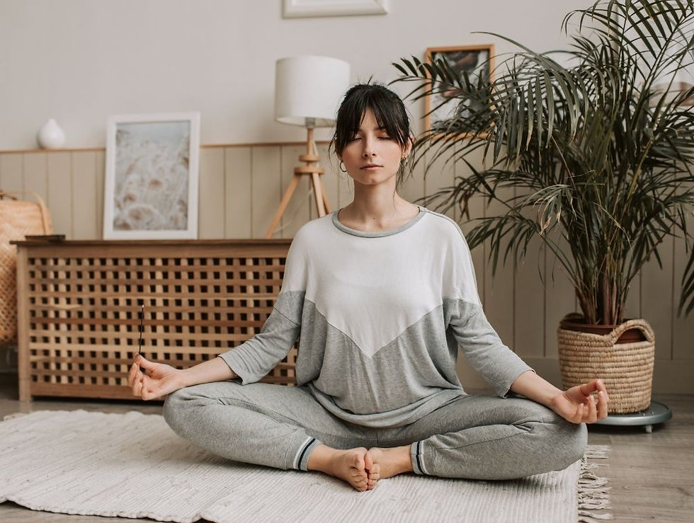 Woman meditating in a peaceful room, seated cross-legged on a white rug. Gray and white attire, surrounded by plants and wooden furniture. Calm mood.