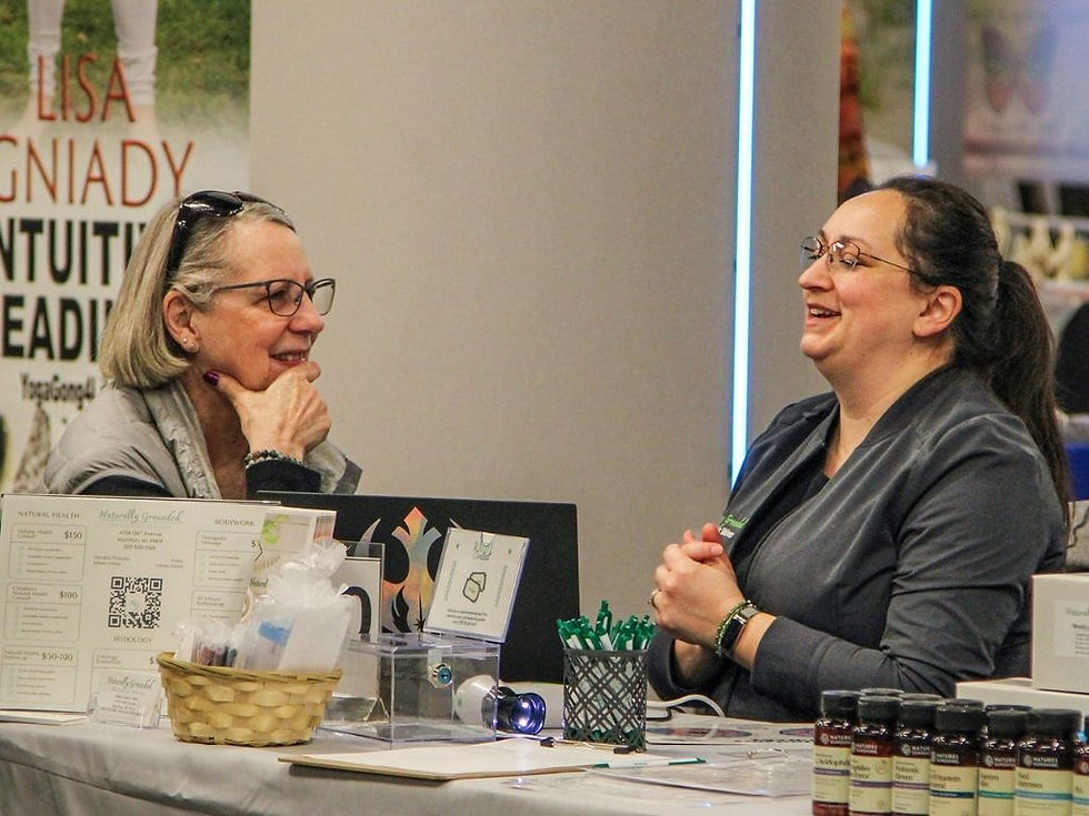 Two women smile and talk at a wellness booth with brochures, pens and product jars; sign mentions Lisa Gniady.