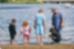 Kids and an adult stand at a lakeshore. One child squats, others look on. A boat is visible in the distance, creating a peaceful vibe.