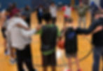 A group of people forms a circle in a gym, arms linked, heads bowed. A basketball lies on the floor, creating a sense of unity and focus.
