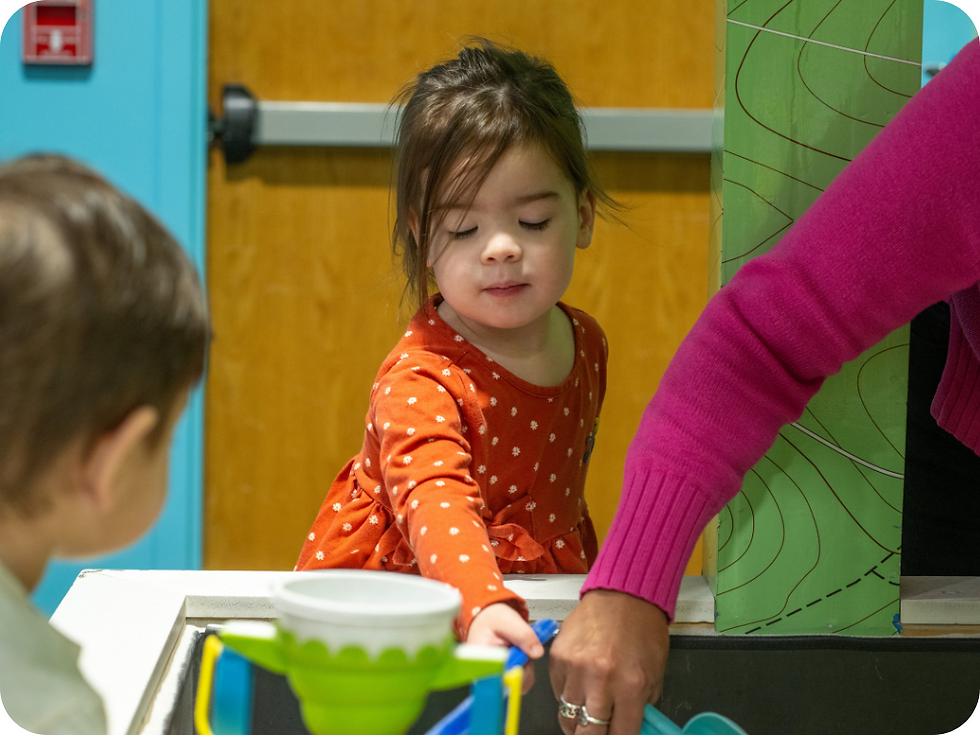 Young girl in an orange polka-dot dress plays with toys on a table, assisted by an adult's hand. Background features a wooden door.