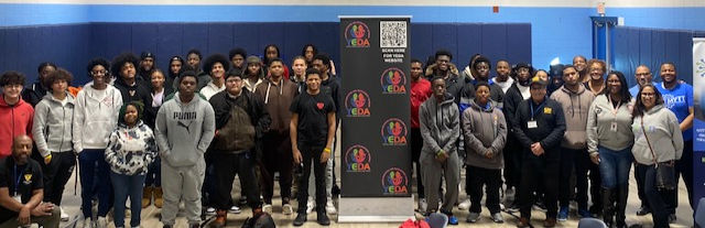 A large group of people poses in front of a "EDA" banner inside a gym. They appear diverse and casually dressed, with a positive mood.