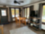 Bright empty dining room with wooden table and chairs, TV, gray curtains, ceiling fan, and large windows overlooking autumn trees