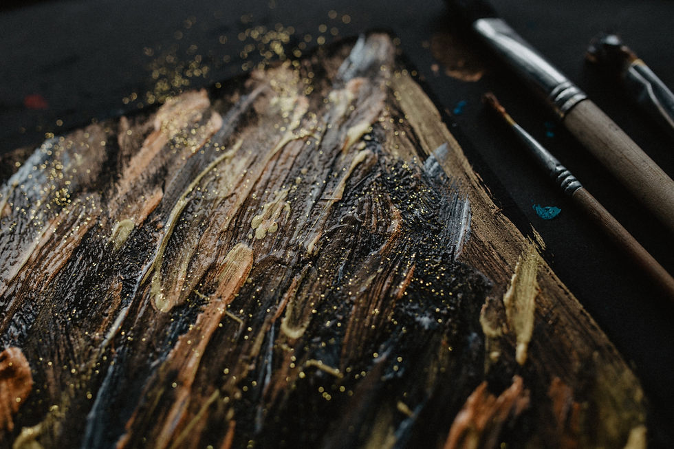 Textured painting with black, gold, and brown swirls. Three paintbrushes lie beside it on a dark surface, with gold specks scattered.
