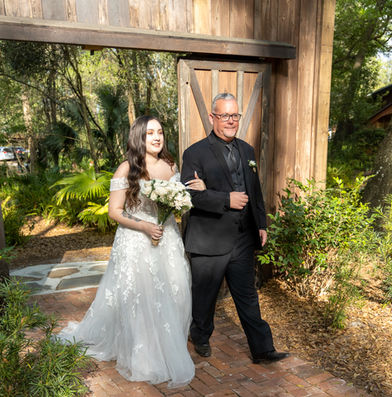 Father of the bride walking her down the aisle. She is in white flower gown with a bouquet of white flowers. He is in all black. 