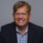 A white man with wavy blonde hair, glasses, and a black suit jacket.