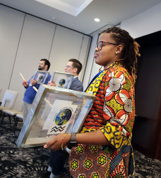 A young woman with dark hair, glasses, and a colorful, patterned shirt holds a square-shaped drum.