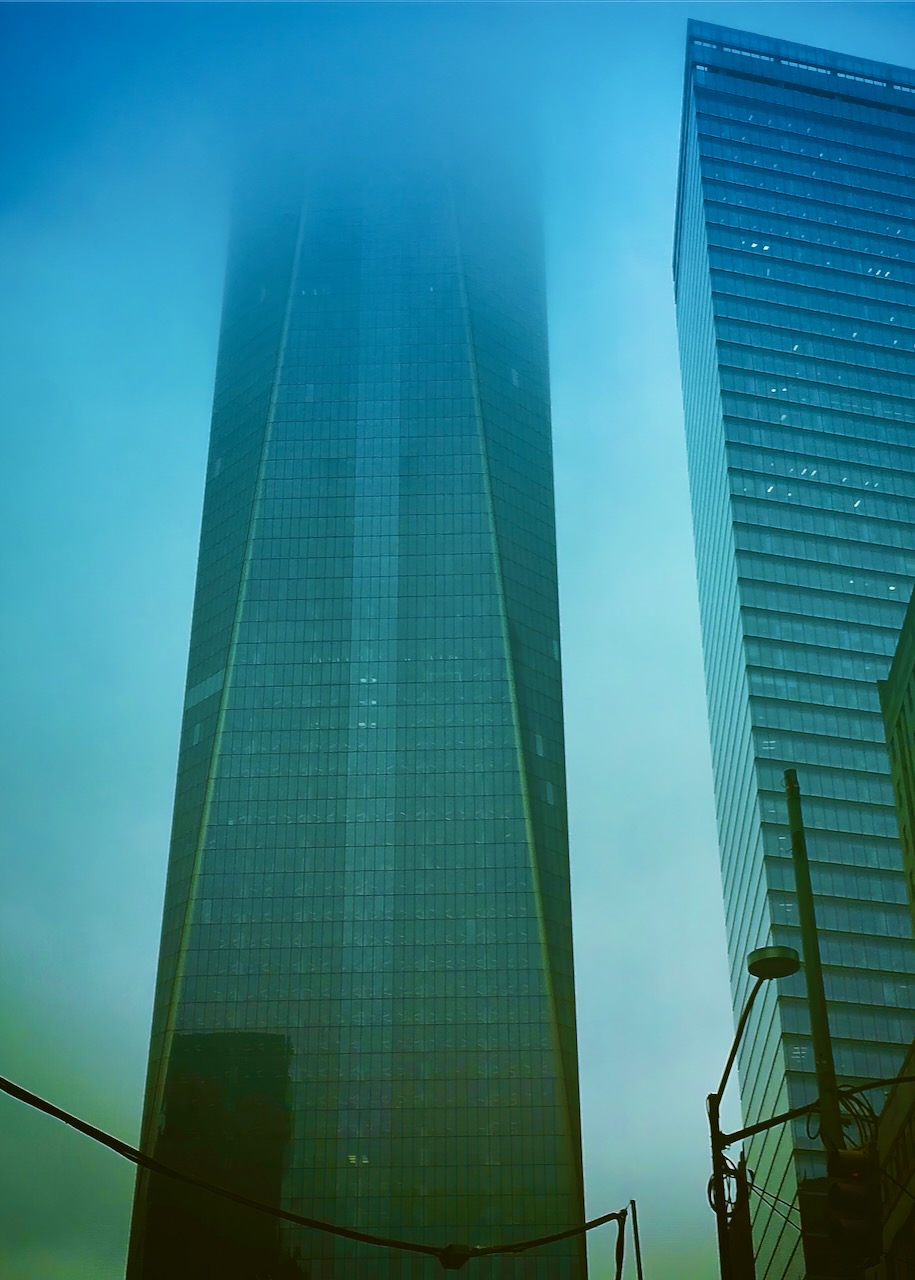 A tall glass tower disappearing into fog beside another high-rise.