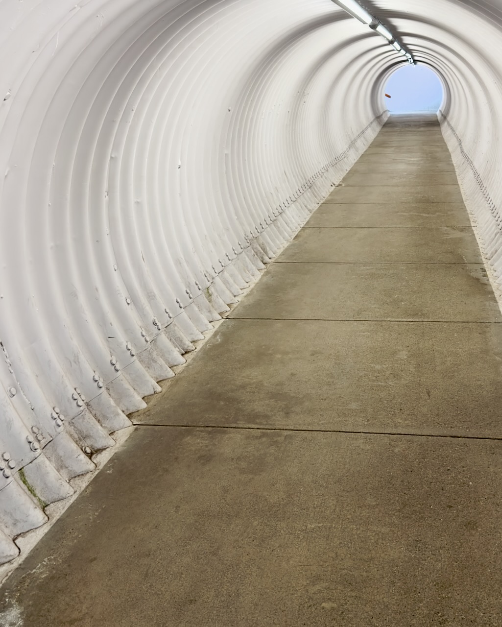 A white ribbed pedestrian tunnel opening toward blue sky.