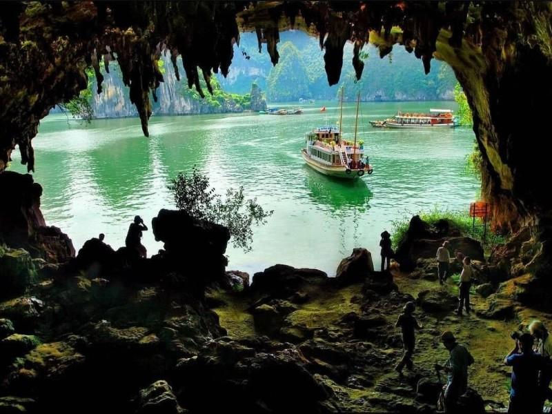 Tourists inside a cave look out at green water and boats in a tropical bay, with jagged stalactites framing the view.