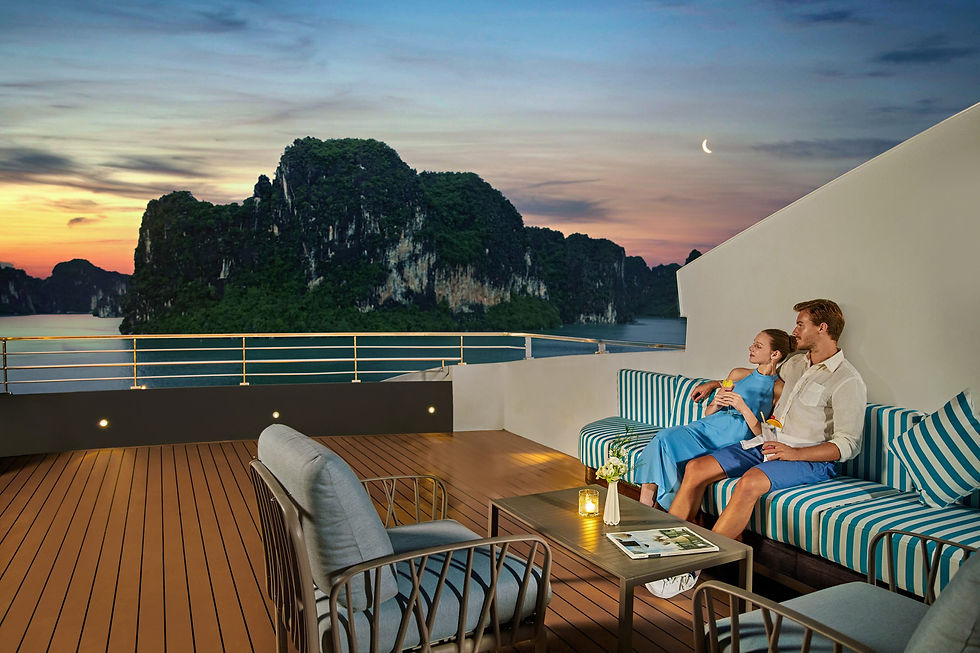 Couple relaxes on a yacht deck at sunset, holding cocktails, with striped blue seating and limestone islands over the sea.