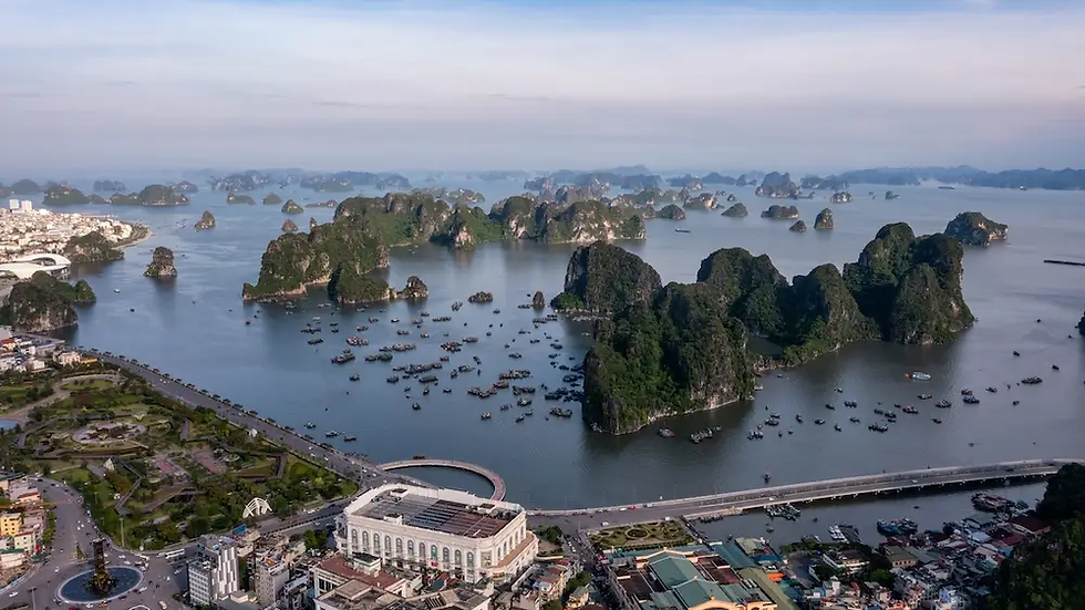 Aerial view of limestone islands and many boats in a calm bay beside a bridge and city at sunrise.