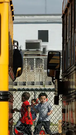 Two school buses parked side by side, with a view of children playing behind a fence.