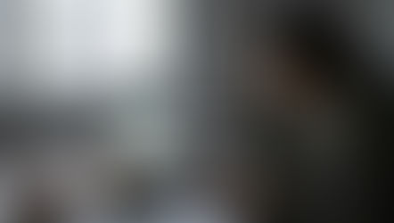 A professional woman sitting at her office desk, resting her head on one hand, staring blankly at her laptop screen. The lighting is slightly dim or greyish (suggesting late afternoon). Next to her is an empty coffee mug and maybe a packet of biscuits.