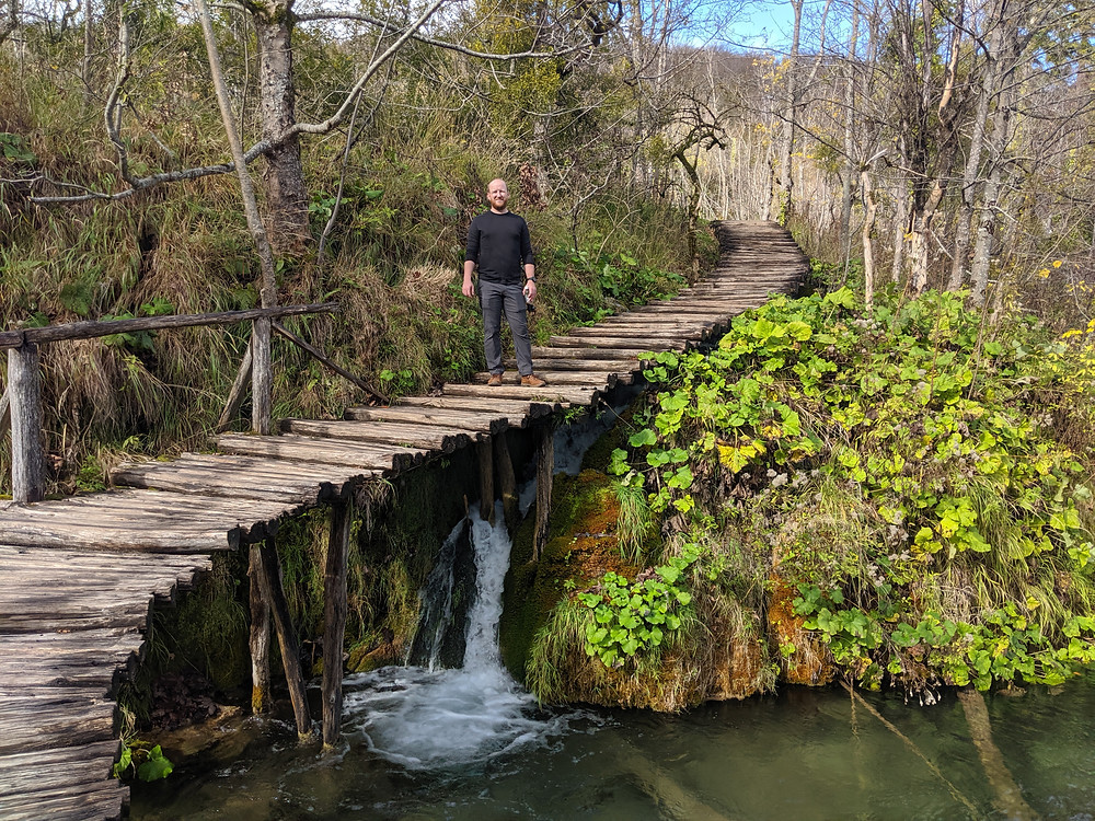 man standing on a boardwalk over a waterfall with lots of greenery surrounding him