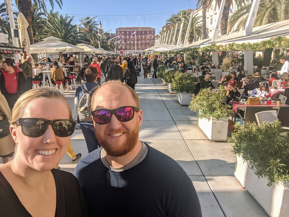 man and woman standing along a busy seaside street with cafes and restaurants in the background