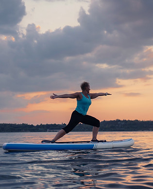 Woman doing the warrior yoga pose on a blue and white paddleboard on the lake at sunset.