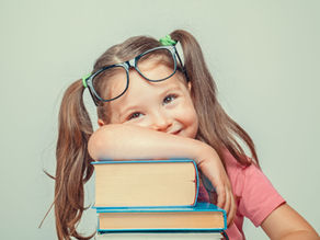 A young girl with red hair and glasses sits cross-legged on the floor reading a book, surrounded by pencils and notebooks. Text overlay highlights a literacy message: "You can never spoil your child with too many books," citing research linking early reading for pleasure with later cognitive and mental health benefits (Sun & Sahakian et al., 2023).