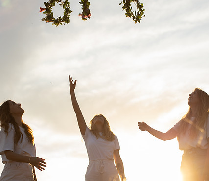 Drei Frauen stehen im Sonnenuntergang und werfen Blumenkränze in die Luft - Symbol für Neubeginn und Verbindung zur Natur