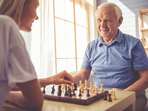 Older man in a blue shirt and a woman playing chess in a sunlit room. The man smiles, creating a warm, relaxed atmosphere.
