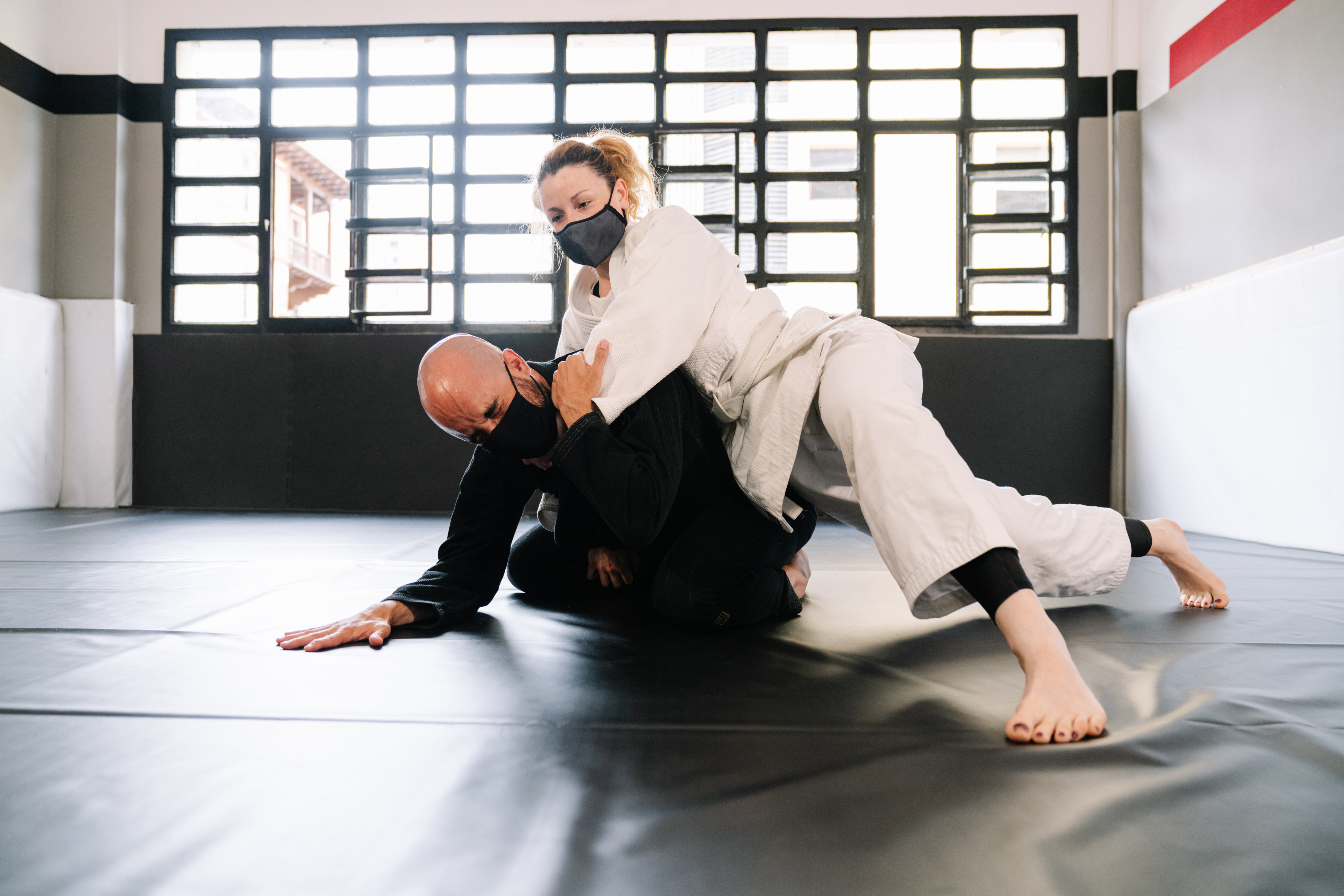Two people in a martial arts studio practicing grappling moves, dressed in traditional uniforms and wearing masks.