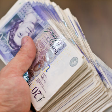 A hand holding a large stack of £20 British banknotes with purple and white tones, featuring text and a portrait, blurred background.