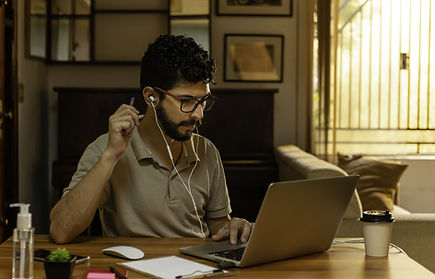 Hombre joven estudiando en la mesa de su sala-comedor al atardecer, con una taza de café, laptop, lentes y audífonos, concent