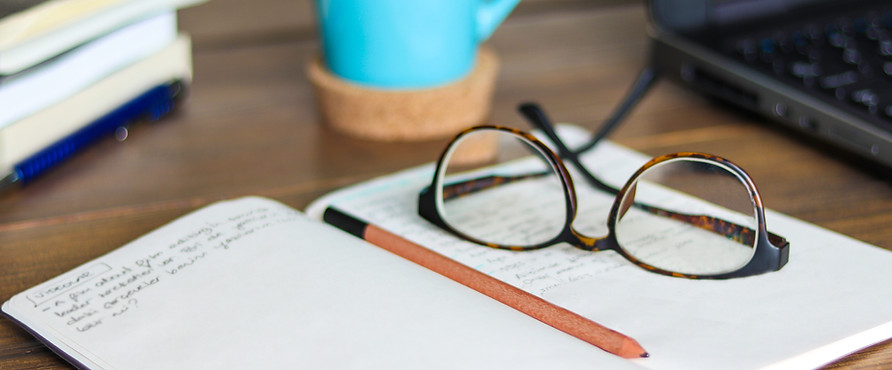 A note book lying open with glasses and pencil resting on it, beside a laptop and mug.