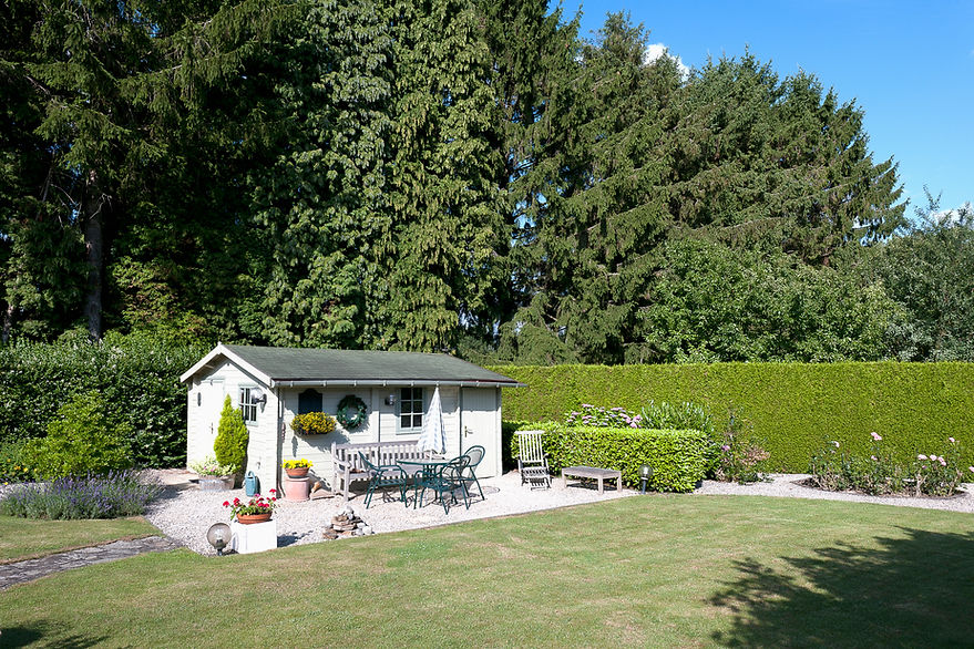 A quaint garden shed with a green roof sits on a patio with outdoor furniture