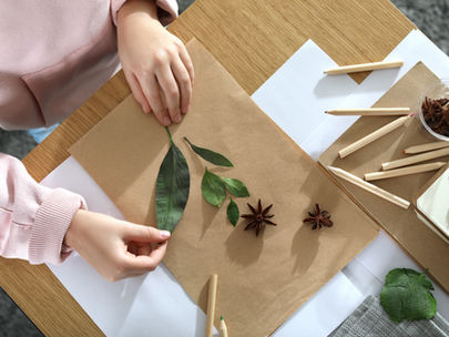 Hands arranging leaves and star anise on brown paper, surrounded by colored pencils on a wooden table. Soft natural light.