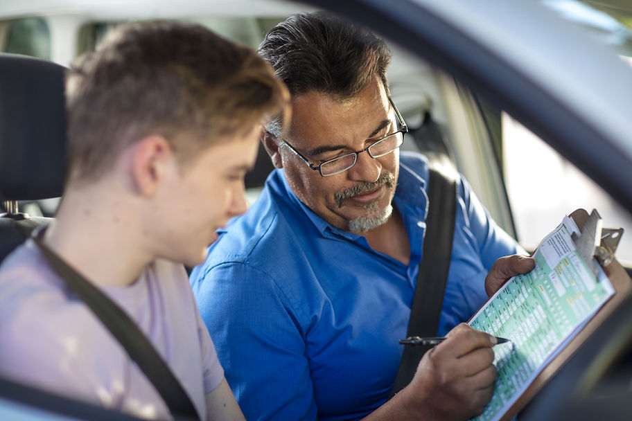 A driving instructor in a blue shirt helps a teenager in a car, reviewing a checklist on a clipboard.