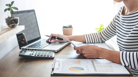 Person in striped shirt works at a laptop with calculator and charts on desk, beside coffee and a potted plant, focused and busy