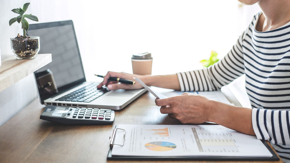 Person working at a desk with a laptop, holding a paper and pen. A calculator, clipboard with charts, coffee cup, and plant are nearby.
