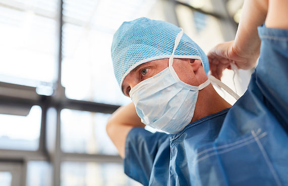 A male surgeon in a blue surgical cap and mask is getting ready for an operation. He is wearing blue scrubs and is standing in a brightly lit room.