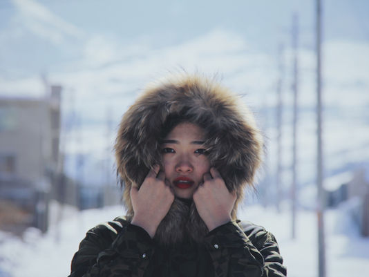 Woman bundled in a winter coat with a fur lined hood, holding the hood close to her face in cold weather, standing outdoors on a snowy city street.