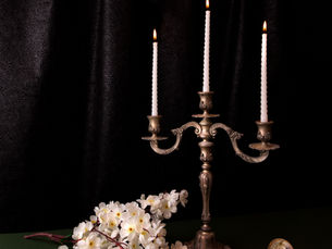 Candles in a brass holder are lit, surrounded by white flowers, pearls, and a compact on a green surface against a dark draped background.
