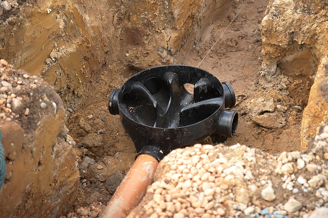 A black circular drainage pipe junction is partially buried in a trench