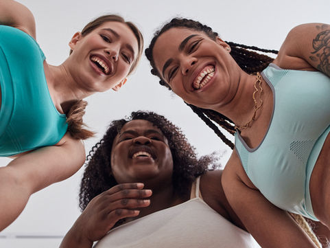 Three women in sportswear smiling and laughing, viewed from below against a white background, conveying joy and friendship.