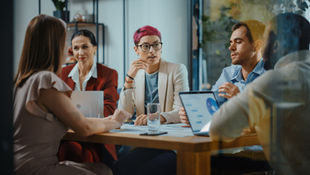 Five people in a meeting room, discussing while looking at laptops and documents. A woman with pink hair speaks. Office setting, focused atmosphere.