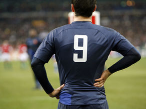 A rugby player is facing the field. The image shows his back with his hand on his lower back