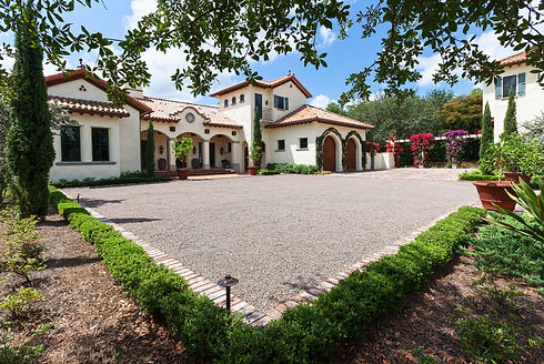 Gravel driveway leading to a large Mediterranean style house with manicured hedges