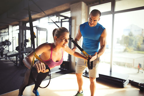 A woman performs suspension training in a gym, smiling with effort, assisted by a man