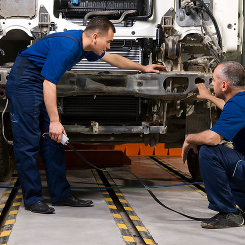 Two mechanics in blue uniforms work on a vehicle chassis in a garage, focusing intently