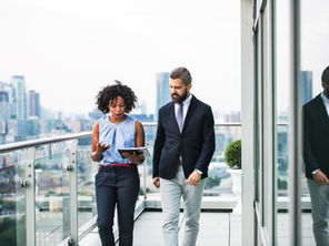 Two businesspeople walk on a rooftop balcony, woman holding a tablet and talking, with a city skyline and glass reflection behind them.