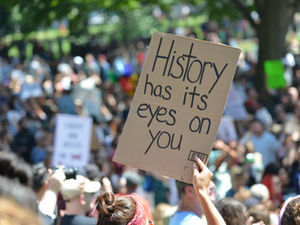 A protester holds a powerful sign reading "History has its eyes on you" amidst a large, engaged crowd, emphasizing accountability and change.