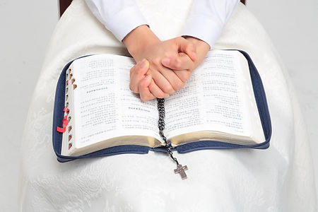 Hands clasped in prayer over an open Korean Bible, with a rosary and cross. The person wears white, creating a peaceful, reverent mood.