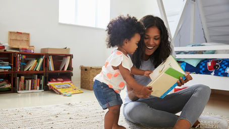 Mère souriante lisant un livre avec sa petite fille dans une chambre lumineuse, sur un tapis beige.