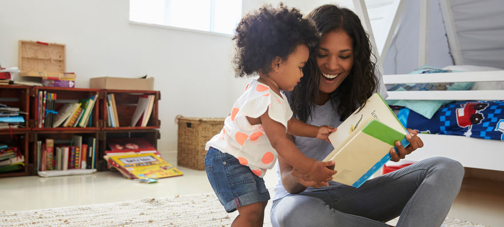 A mom and a child are sitting together on the floor reading a book together. They are in a bright room with a bookshelf behind them that has many books on it.